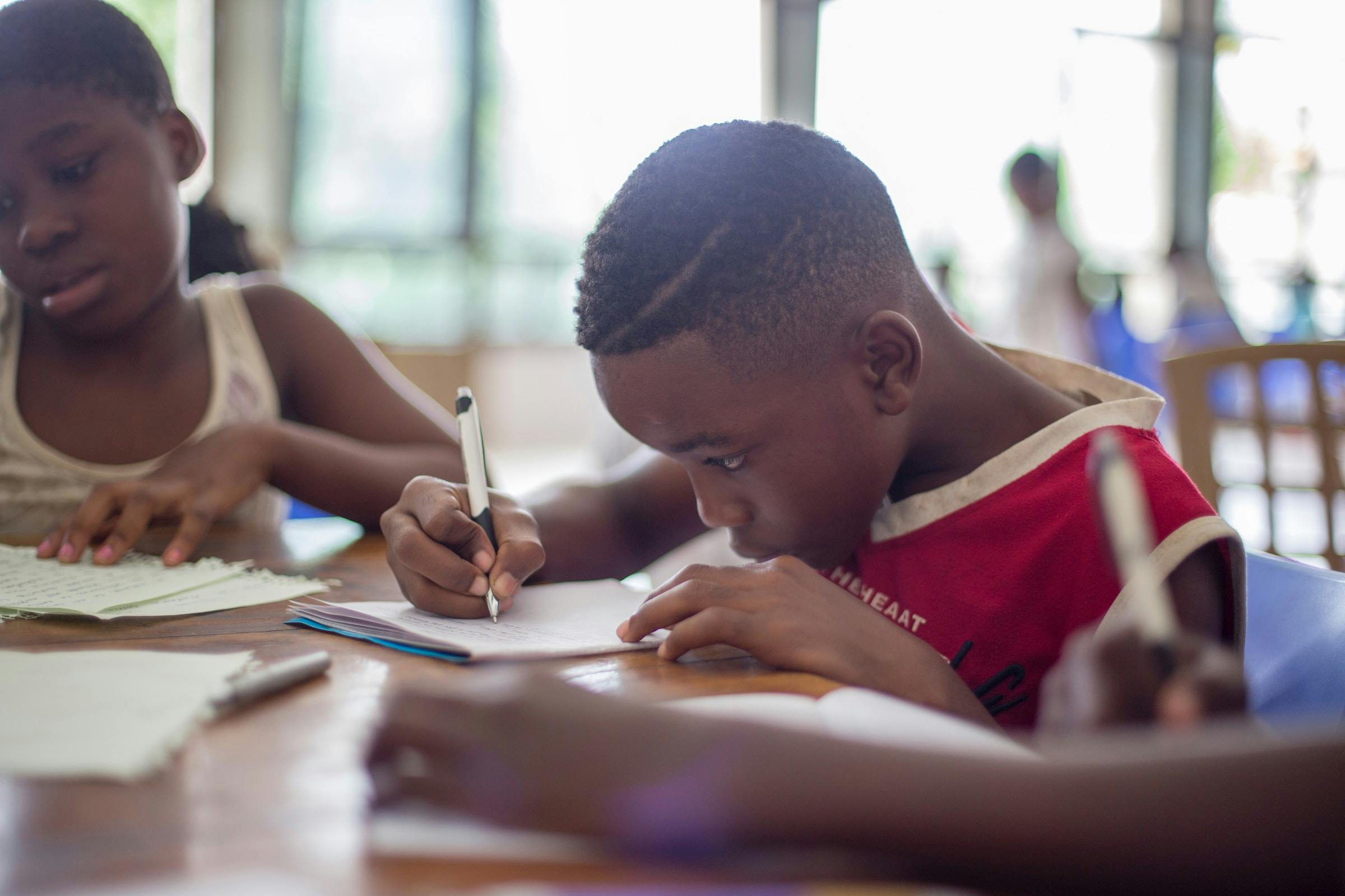 Boy writing on paper near girl in a school-like setting.